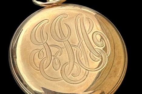 A gold pocket watch with engraved lettersDescription automatically generated